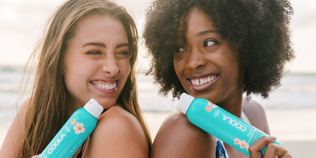 women standing back to back holding COOLA Sunscreen Spray
